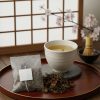 和室の盆に載った湯呑みと、手前に置かれたテトラ型の阿波晩茶ティーバッグ、そして乾燥した茶葉。背景には障子と桜の花が見える。