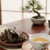 湯呑に注がれた阿波晩茶と乾燥した茶葉が皿に盛られ、後方に鉄瓶と盆栽が置かれた和室の情景。